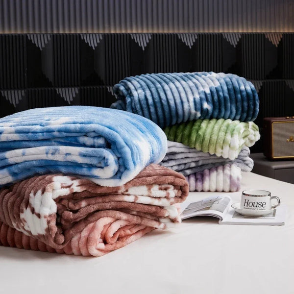 A stack of tie-dye flannel fleece throw blankets in shades of blue, green, and brown are displayed on a white surface.