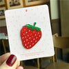 A white card featuring a red strawberry embroidery patch, with a hand holding the card in a blurred indoor setting.