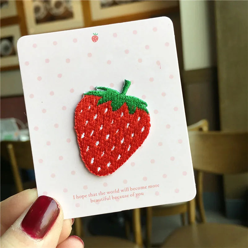 A white card featuring a red strawberry embroidery patch, with a hand holding the card in a blurred indoor setting.