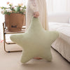 A light green star-shaped plush throw pillow resting on a carpet next to a white bed and a potted plant.
