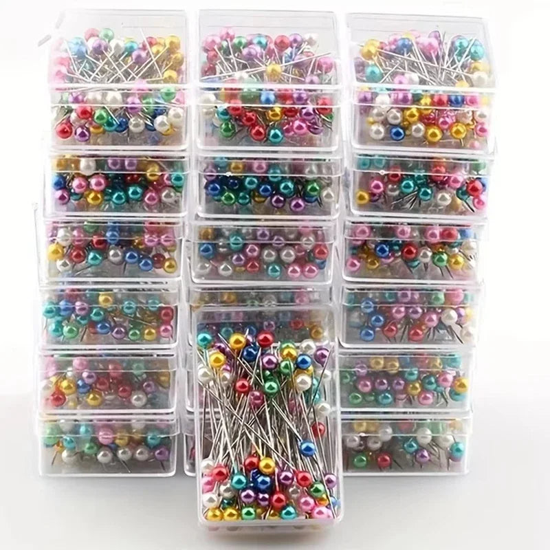 A collection of decorative sewing and pinning needles in various colors and styles packaged in clear plastic containers.
