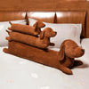 A stack of three brown corduroy dachshund body pillows resting on a white bed with light floral accents.