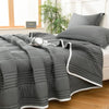 A gray quilted blanket is draped over a bed, next to a window with greenery.