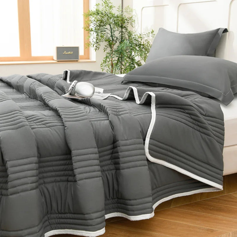 A gray quilted blanket is draped over a bed, next to a window with greenery.