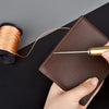 A hand holds a brown leather sewing awl and stitching kit next to a spool of golden thread.