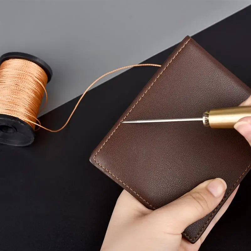 A hand holds a brown leather sewing awl and stitching kit next to a spool of golden thread.