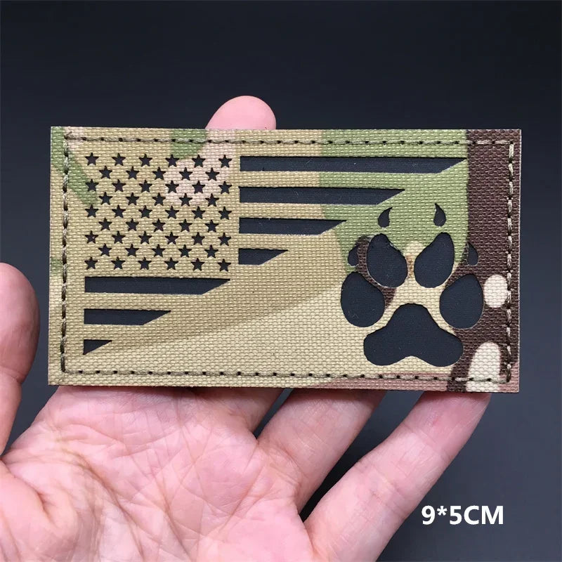 A rectangular tactical patch featuring a camouflage design with American flag elements and paw prints.