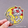 Yellow embroidered patch featuring a white skull with red crosses, showing the detail of skeletal hands.