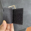 A close-up of a black tactical hook and loop patch with a brass hook, held by a hand.