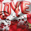 Red 3D letter balloons spelling LOVE scattered on white fabric with red rose petals