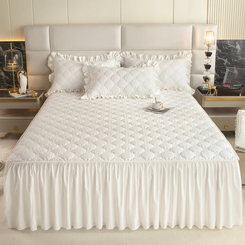 A white quilted bed skirt cover with a ruffled edge and white pillows on a bed.