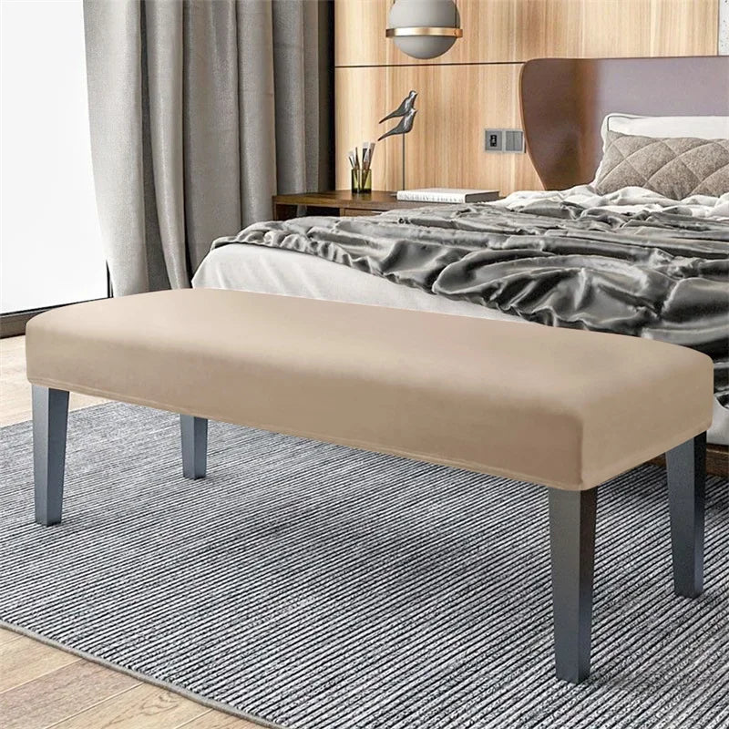 A beige velvet bench slipcover with secure straps placed on a gray striped rug in a bedroom setting.