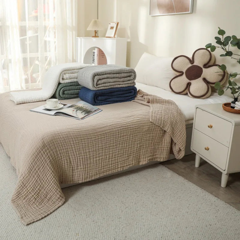 A beige cotton gauze blanket is draped over a bed, with stacked blue and gray towels nearby.