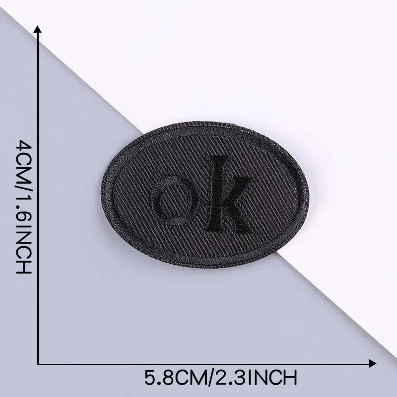 A black embroidered leather patch with the letter OK on it, shown against a gray background.