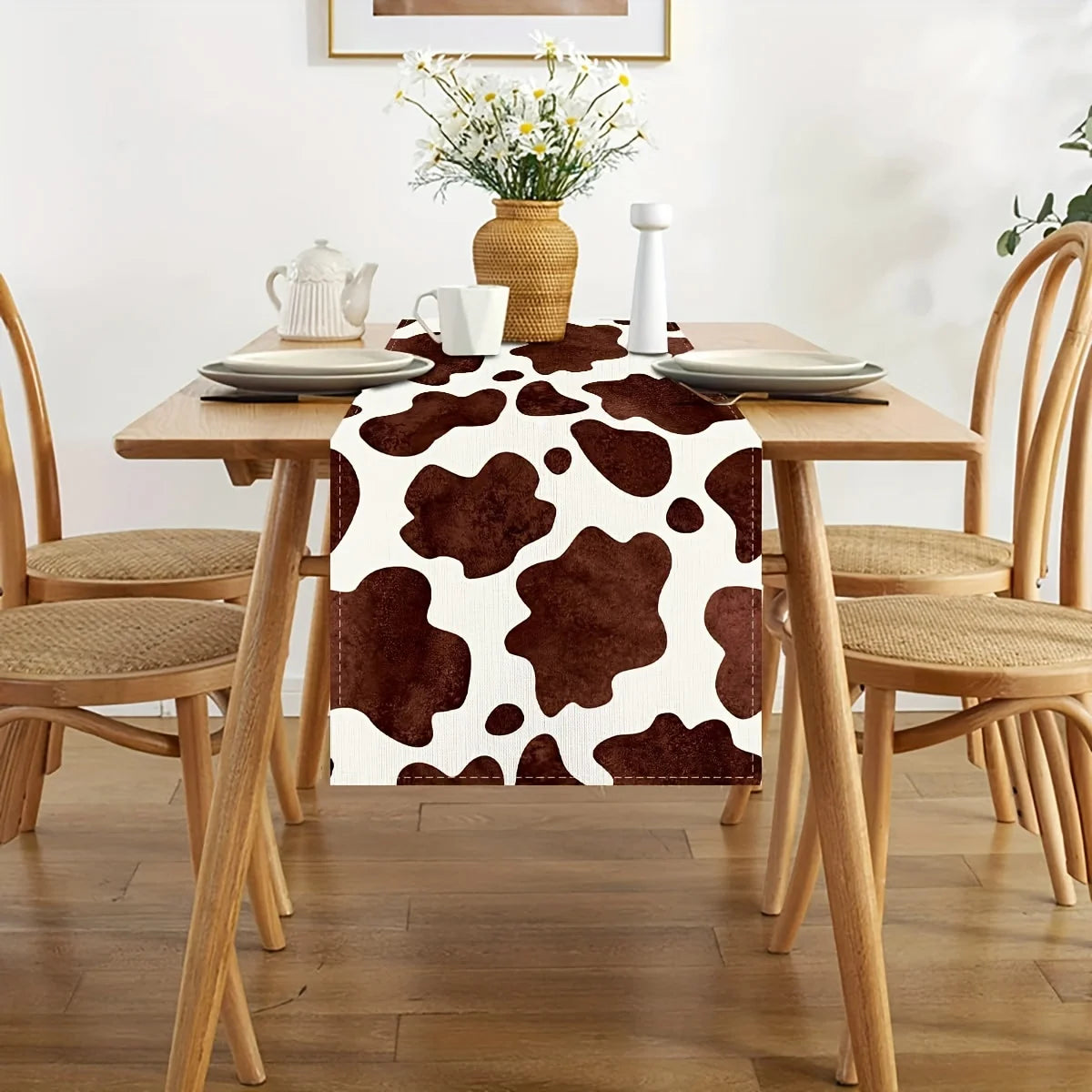 A brown cow print linen table runner displayed on a wooden dining table with white dishes and floral decor.