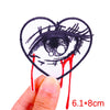 A hand holds up a heart-shaped embroidered patch depicting an eye with a tear running down it.