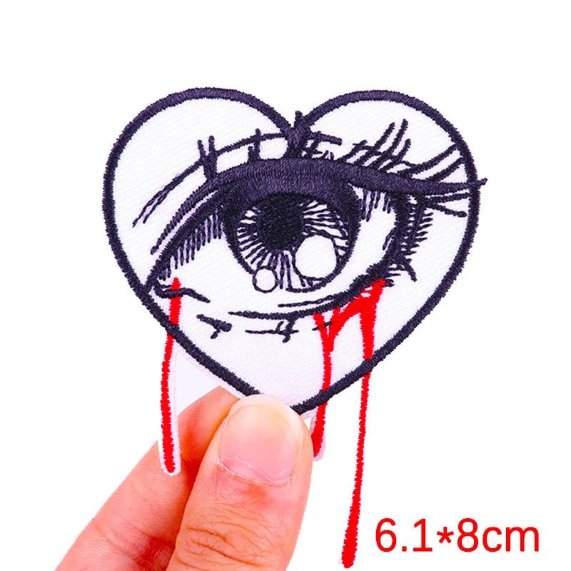 A hand holds up a heart-shaped embroidered patch depicting an eye with a tear running down it.