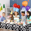 A table set for a birthday party featuring a cow print disposable plastic tablecloth and colorful balloons and cake