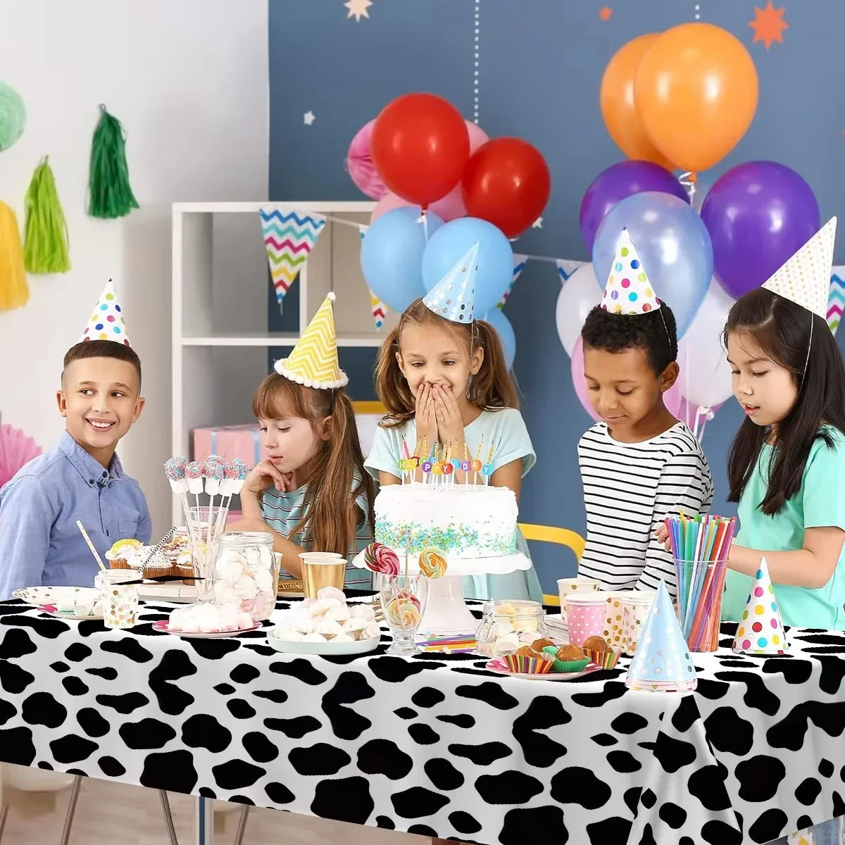 A table set for a birthday party featuring a cow print disposable plastic tablecloth and colorful balloons and cake