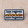 An enamel pin featuring the text less regrets more baguettes on a rectangular background.