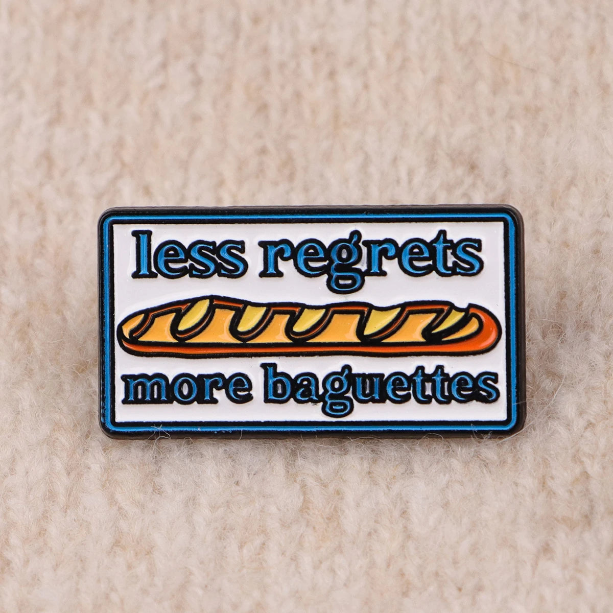 An enamel pin featuring the text less regrets more baguettes on a rectangular background.