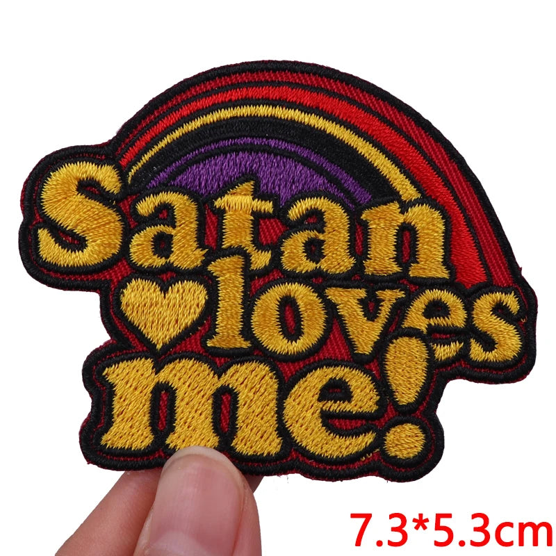An embroidered patch shaped like a devil's head with the text Satan loves me in yellow and red thread.