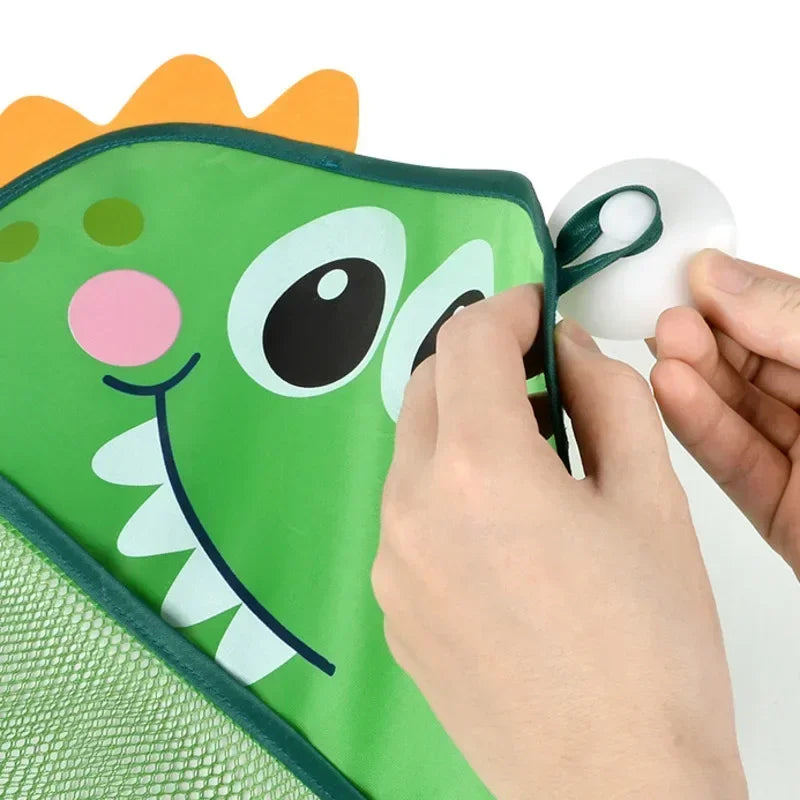 A hand is opening a green dinosaur-themed bath toy organizer bag with a mesh net and a white silicone bath toy.