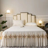 A luxurious white bed with embroidered cotton bedspreads and pillows against a light gray wall.