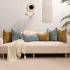 A beige sofa decorated with various textured throw pillows and a cream knitted blanket against a white wall.