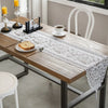 A wooden table is set with a white lace table runner featuring black embroidery and gold tassels, alongside tea service and d