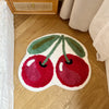 A soft tufted area rug shaped like a green apple with red cherries on a light wood floor.