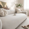 A white quilted sofa cover with elastic straps is displayed on a light-colored sofa in a bright room.
