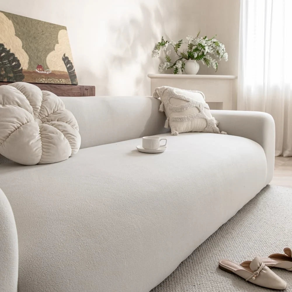 A white quilted sofa cover with elastic straps is displayed on a light-colored sofa in a bright room.