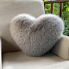 A fluffy gray heart-shaped fox fur throw pillow resting on a light-colored sofa.