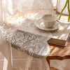 A sheer floral lace curtain draped over a table, featuring intricate white embroidery and beige fabric.