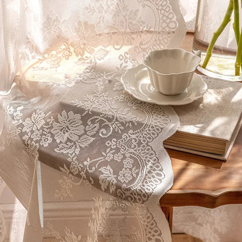 A sheer floral lace curtain draped over a table, featuring intricate white embroidery and beige fabric.