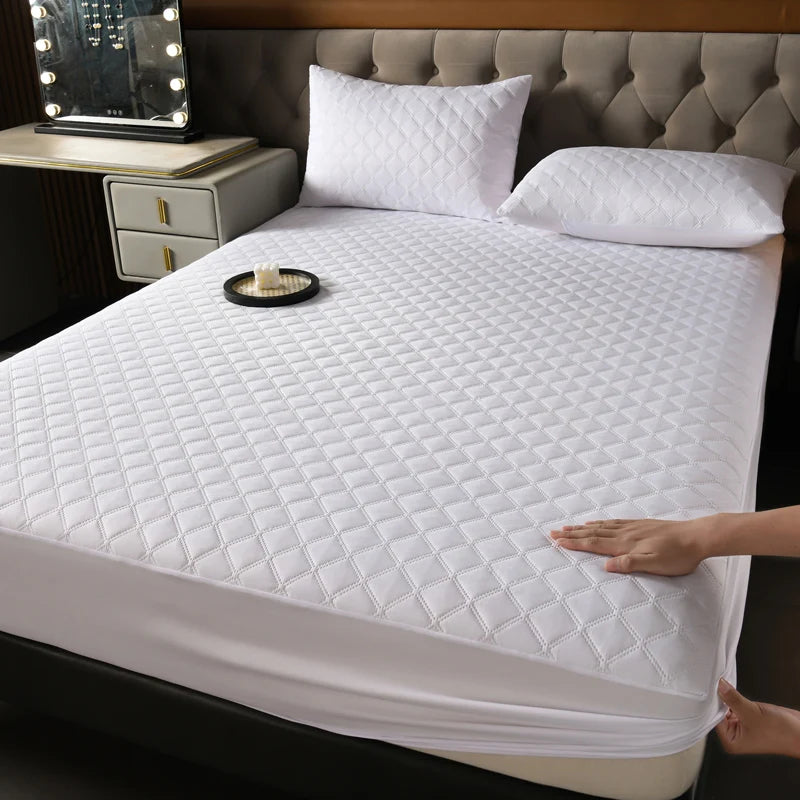A white quilted mattress protector is shown on a bed, next to a nightstand and pillows.
