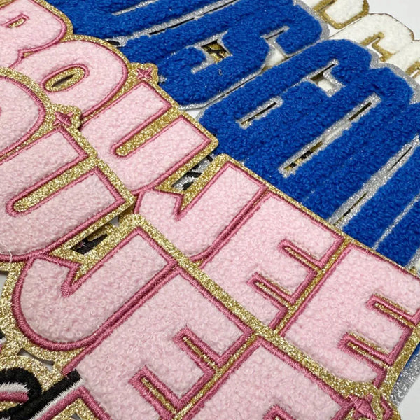 A close-up of three iron-on embroidery patches in pink, blue, and gold thread on a fabric background.
