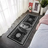 A black decorative floor mat featuring a 100 dollar bill design placed on a gray floor next to a white bed.