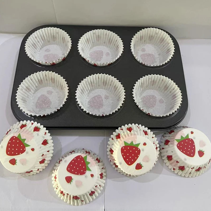 A black baking tray holds twelve white paper cups with a pink strawberry pattern, and four individual cups are displayed belo