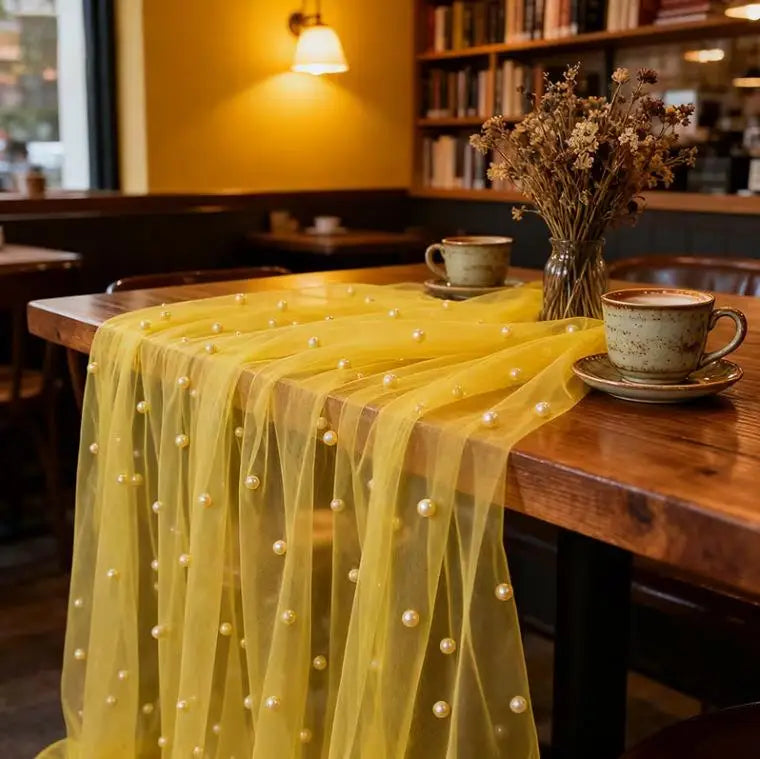 A sheer yellow pearl beaded tulle table runner draped over a wooden table in a cafe setting.
