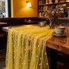 A sheer yellow pearl beaded tulle table runner draped over a wooden table in a cafe setting.