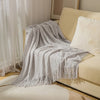 A light gray oversized knitted tassel throw blanket draped over a beige sofa.