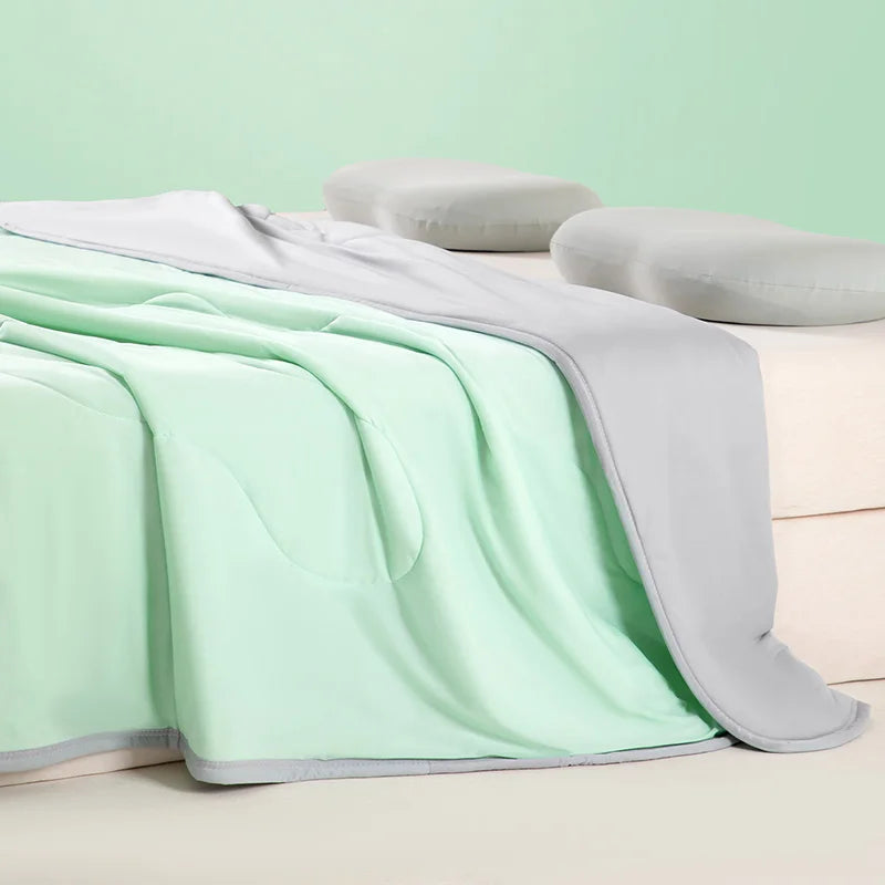 A light mint green ice silk summer cooling blanket draped over a white surface with two gray pillows.