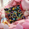 A pink and purple themed square pillow cover with a black background featuring colorful rainbow and floral designs.