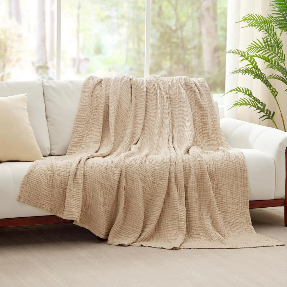 A beige cotton gauze blanket draped over a white sofa near a window.