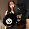 A woman holds a black plush pillow shaped like an eight ball, featuring white markings, against a blurred background.