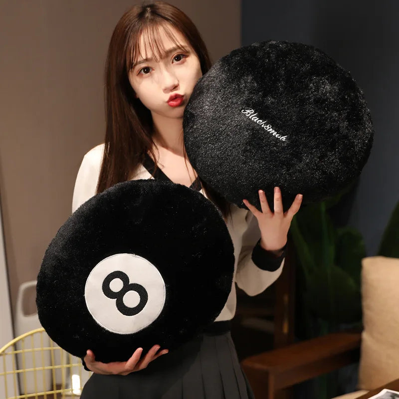 A woman holds a black plush pillow shaped like an eight ball, featuring white markings, against a blurred background.