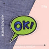 A bright lime green embroidery patch shaped like a speech bubble with the text OK! sewn onto a dark blue fabric.