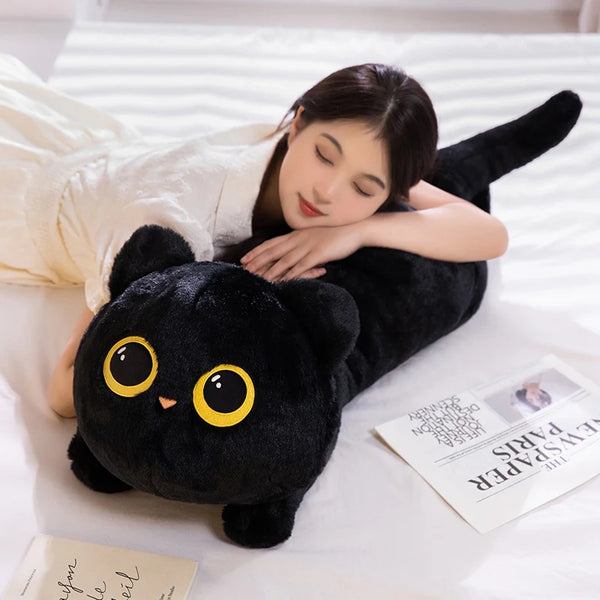 A black cat plush pillow is being held by a person lying down on white bedding, next to a newspaper.